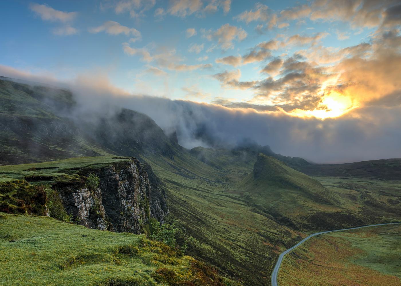 A grassy cliff overlooking a road on the right side of the image, with a cloud sunset in the background.